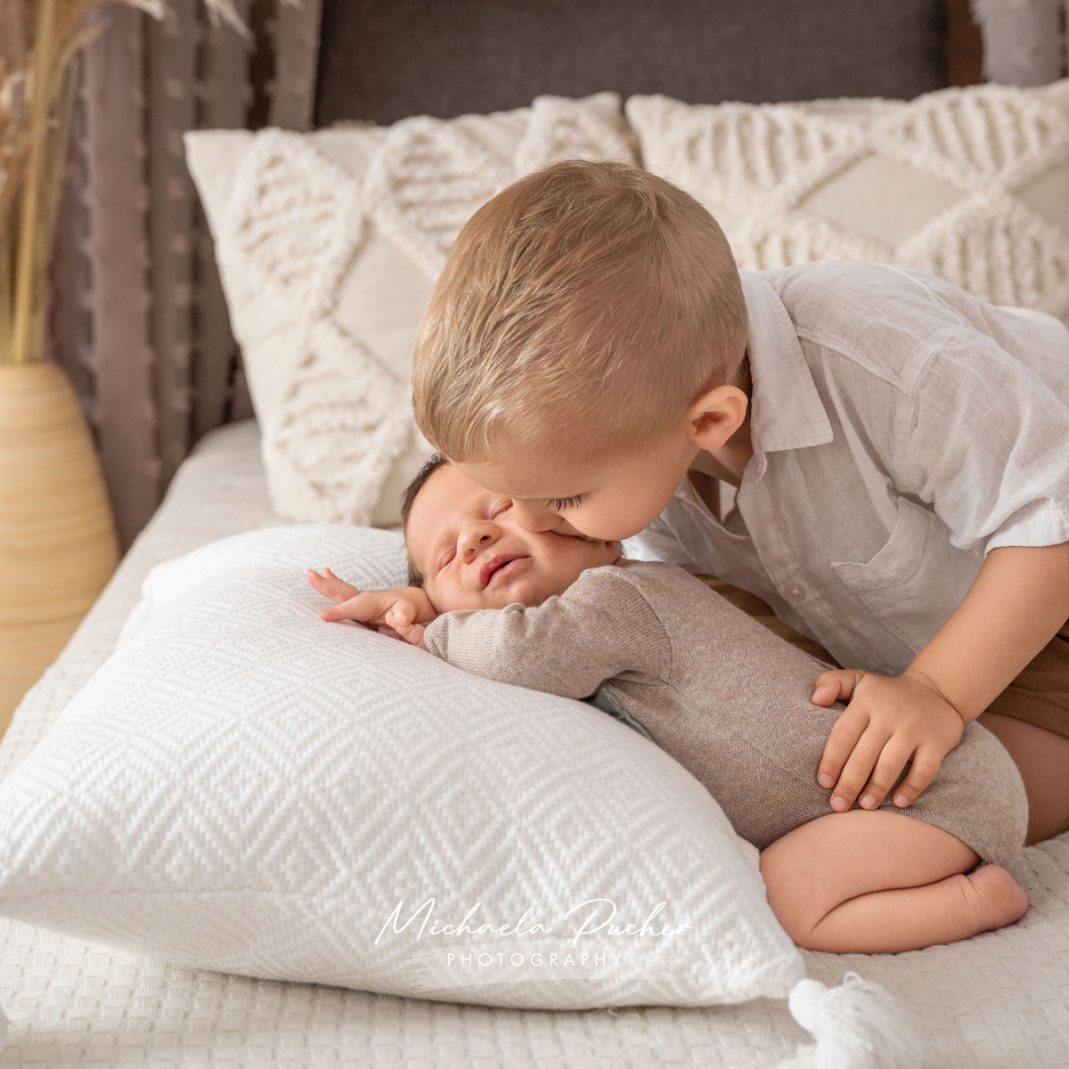 Älteres Geschwisterkind gibt dem lächelnden Baby einen liebevollen Kuss auf die Wange. Beide sitzen auf einem hellen Bett mit weichen Kissen in neutralen Naturtönen.